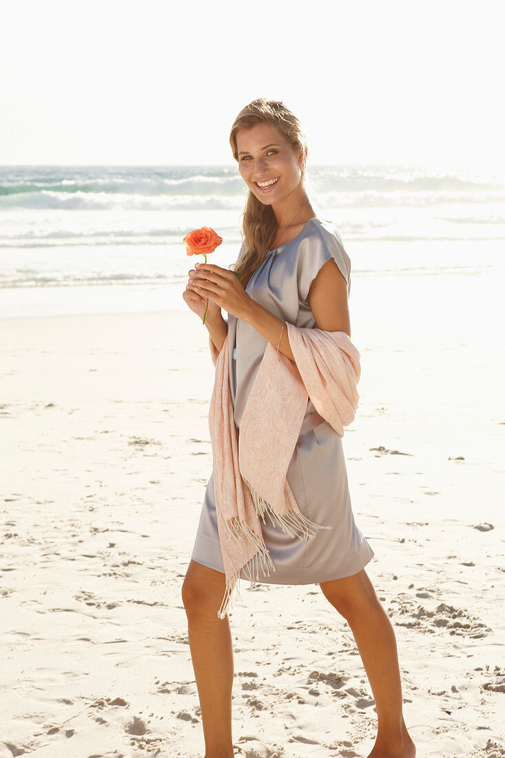 A mature blonde woman on a beach wearing a silver summer dress and holding an orange flower