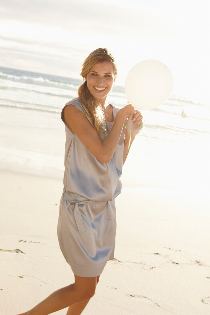 Reife blonde Frau mit silbernem Sommerkleid und weißem Luftballon am Strand