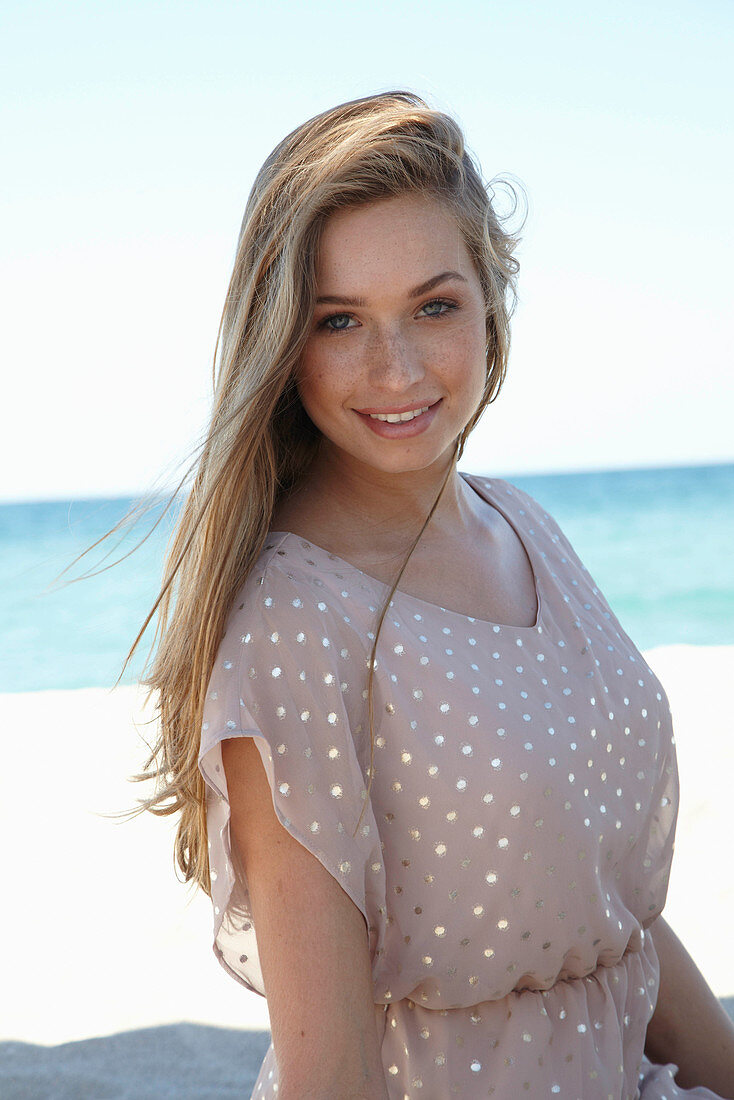 A young blonde woman on a beach wearing a beige polka-dot dress