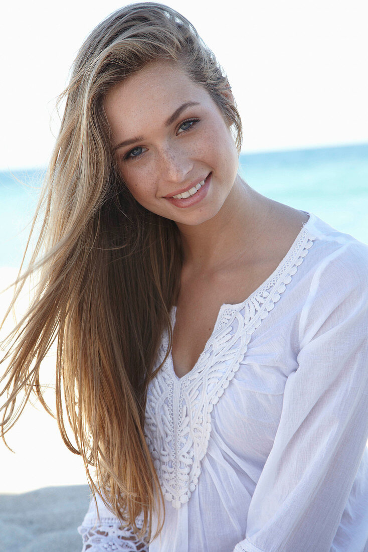 A young blonde woman on a beach wearing a white summer dress