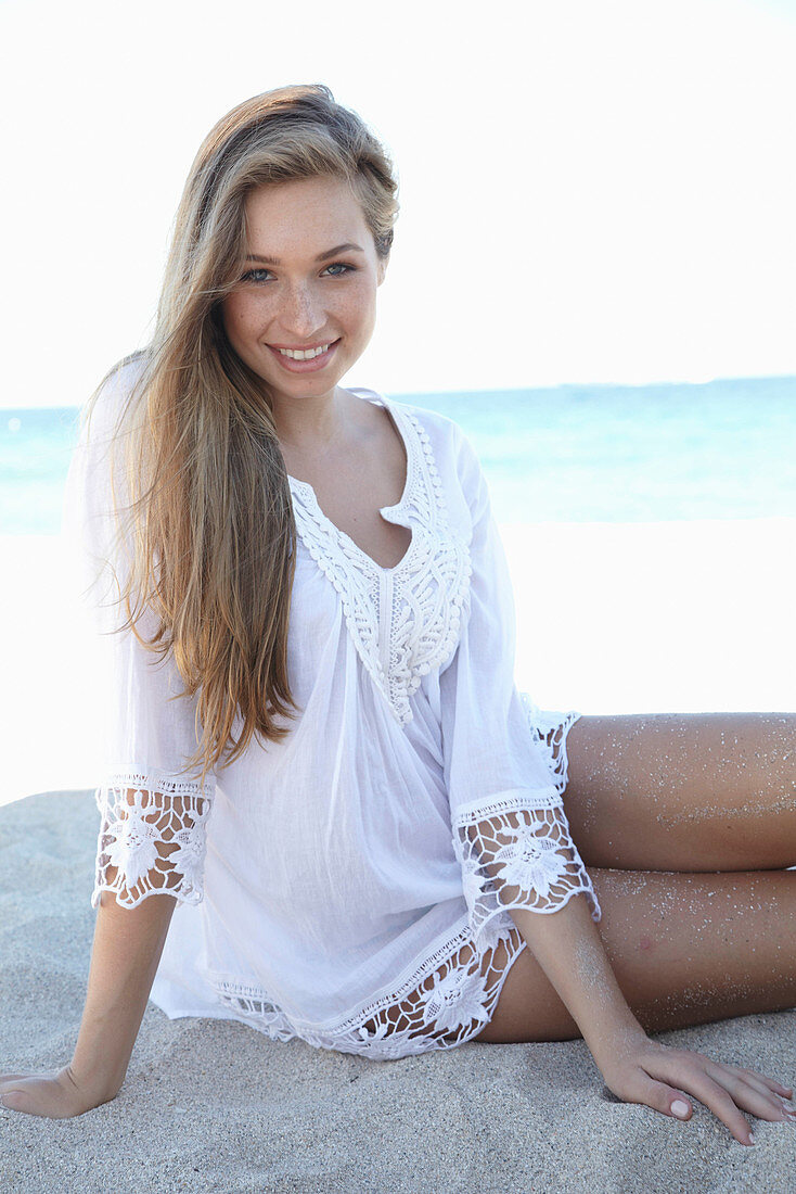 A young blonde woman on a beach wearing a white summer dress