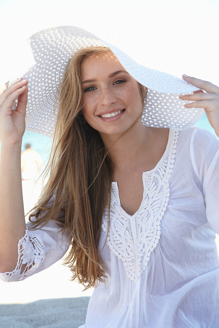 A young blonde woman on a beach wearing a white summer dress and a white summer hat