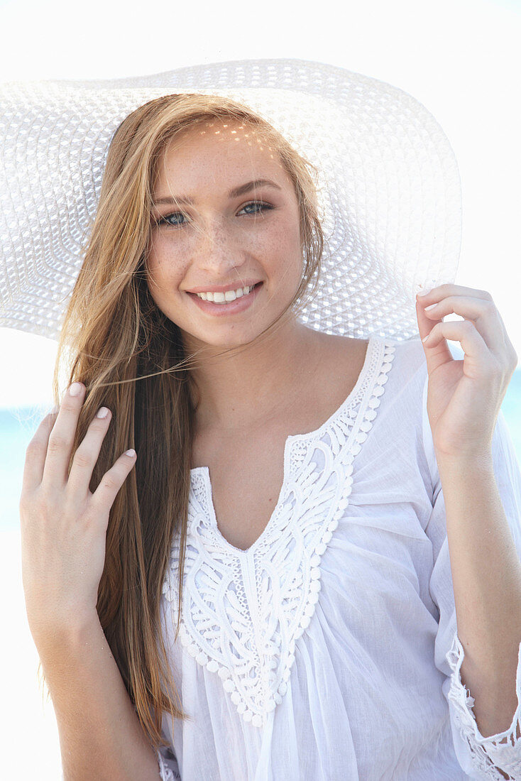 Junge blonde Frau mit weißem Sommerhut in weißem Sommerkleid am Strand