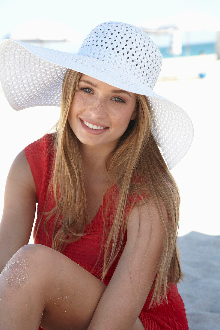 Junge blonde Frau in rotem Sommerkleid mit weißem Hut am Strand