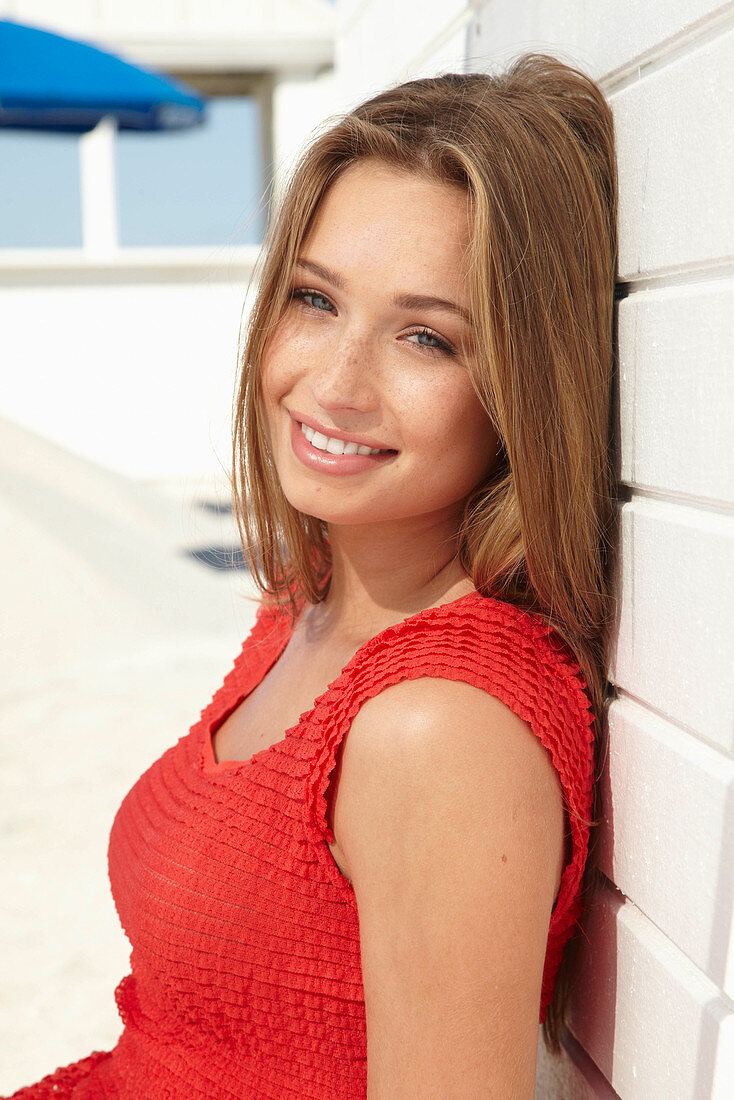A young blonde woman wearing a red summer dress leaning against a wall