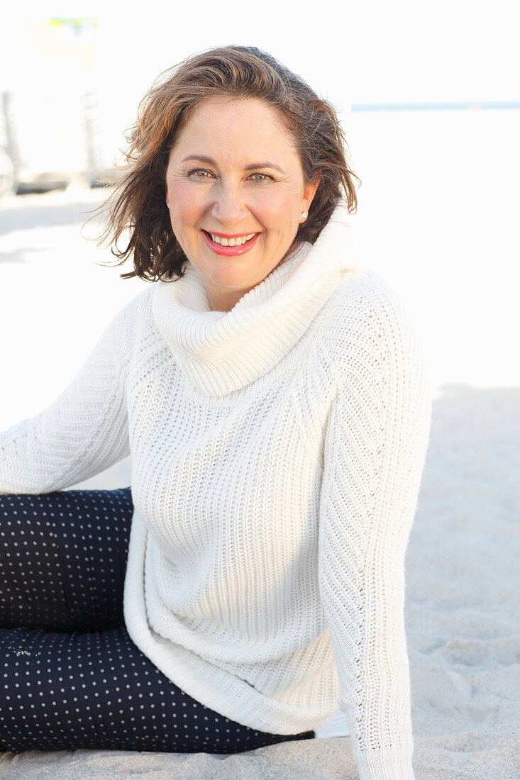 Brünette Frau in weißem Strickpullover und dunkler Hose am Strand