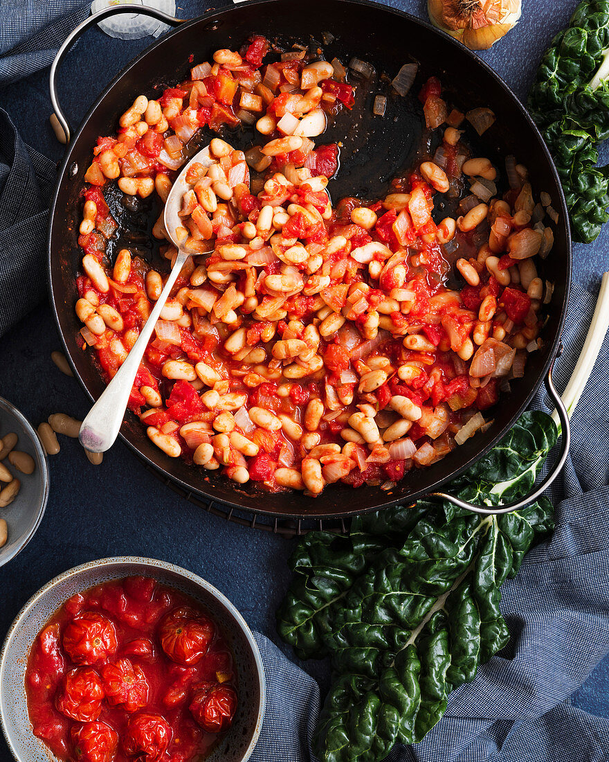 Tomatoes, cannellini beans and onions in a pan beside a bunch of silverbeet and a bowl of cherry tomatoes