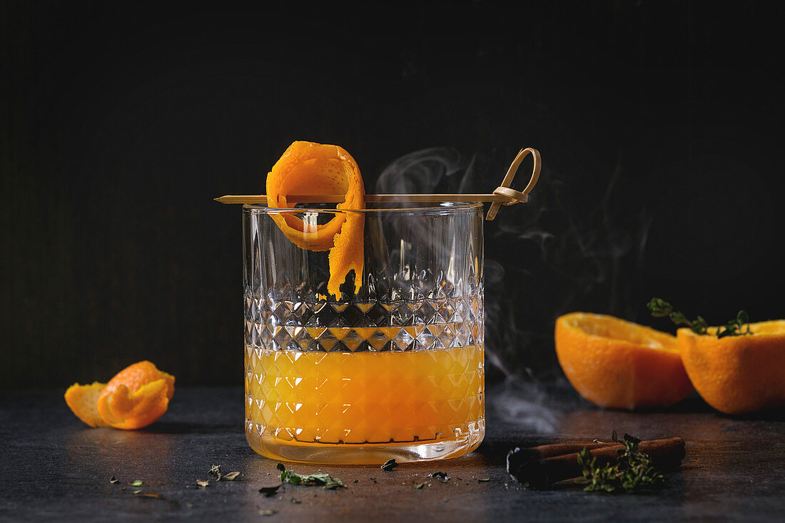 Whiskey mit Orangensaft serviert mit Orangenschalenspieß und rauchender Zimtstange
