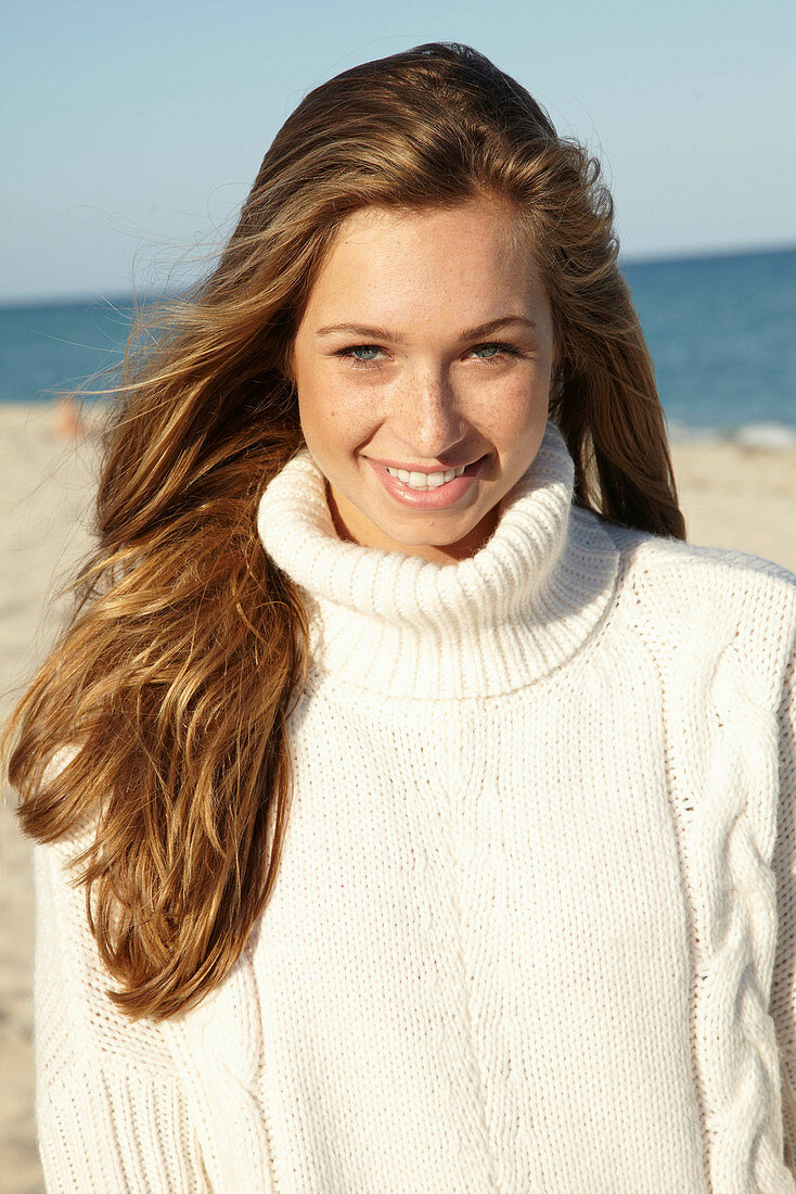 A young blonde woman on a beach wearing a white turtle neck jumper