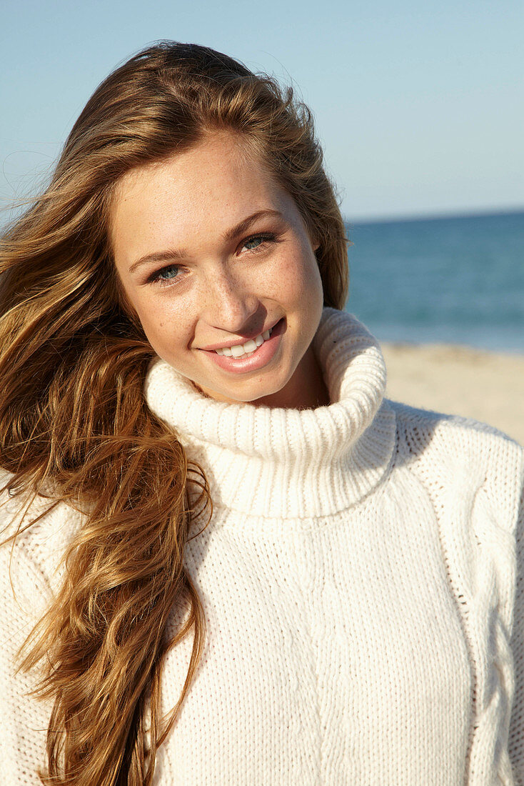 A young blonde woman on a beach wearing a white turtle neck jumper