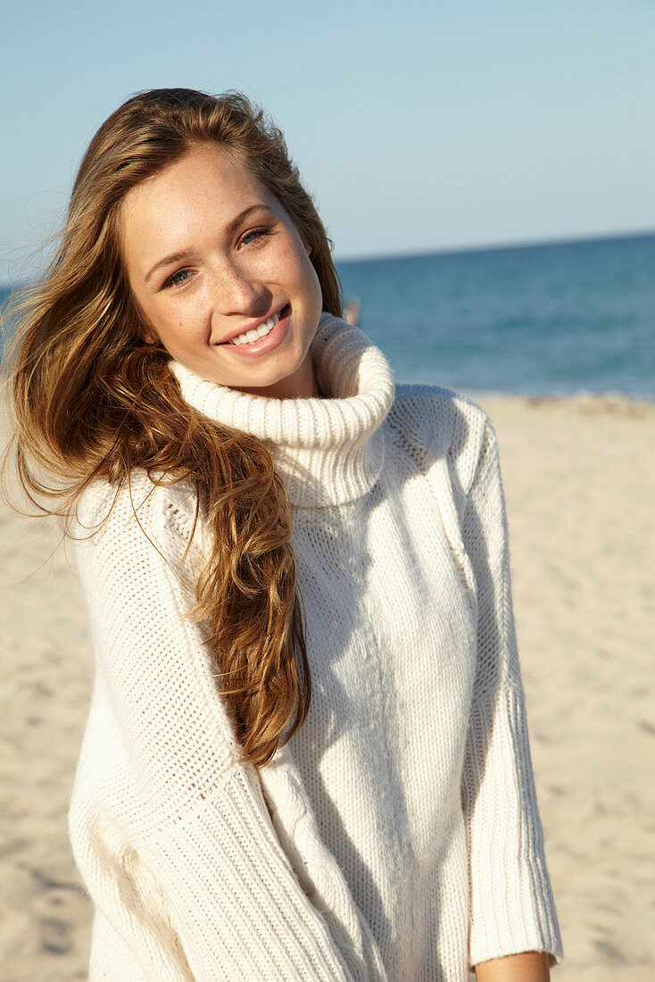 A young blonde woman on a beach wearing a white turtle neck jumper