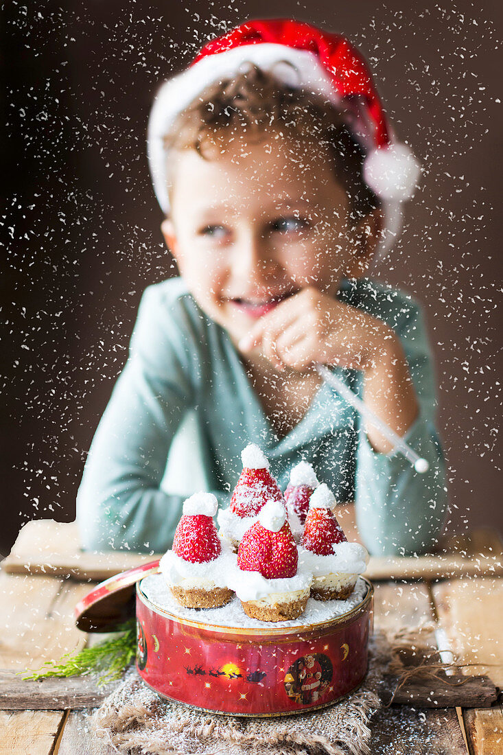 Junge sitzt vor Mini-Käsekuchen in Keksdose dekoriert mit Weihnachtsmützen aus Erdbeeren