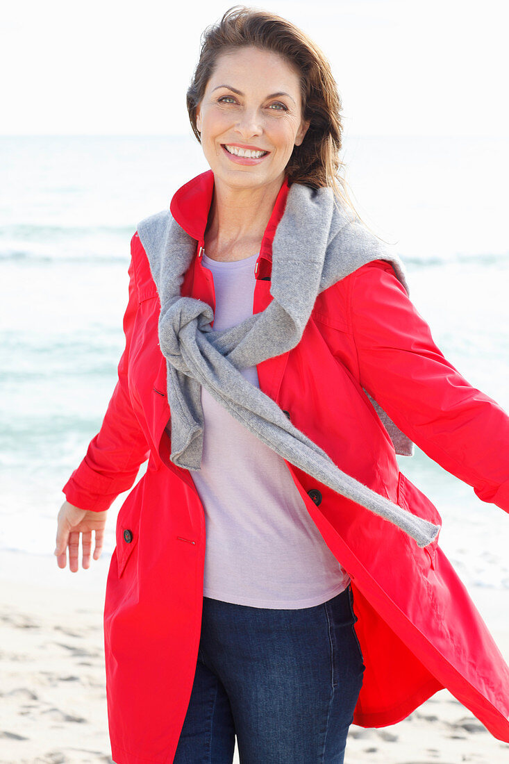 Brünette Frau in rotem Trenchcoat mit grauem Pulli über den Schultern am Strand