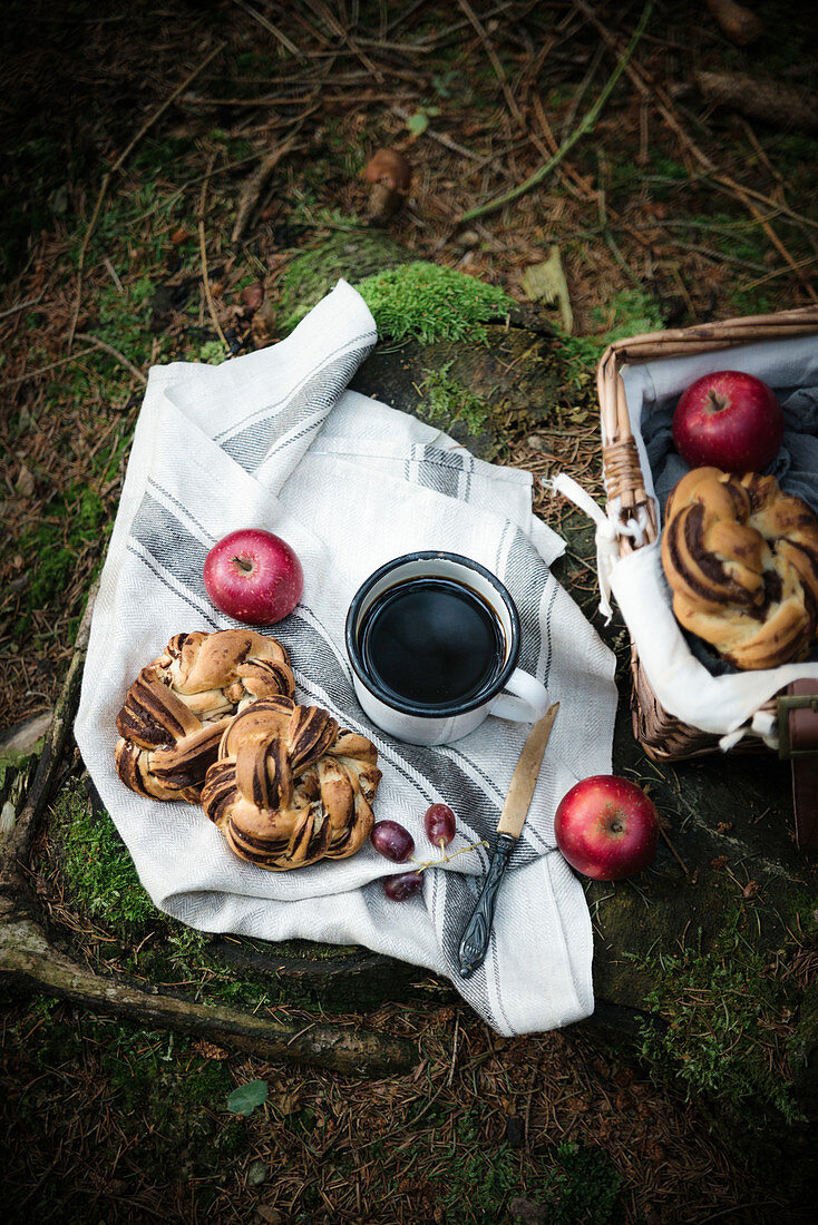 A picnic basket in a forest, filled with a vegan chocolate bun, fruit and coffee