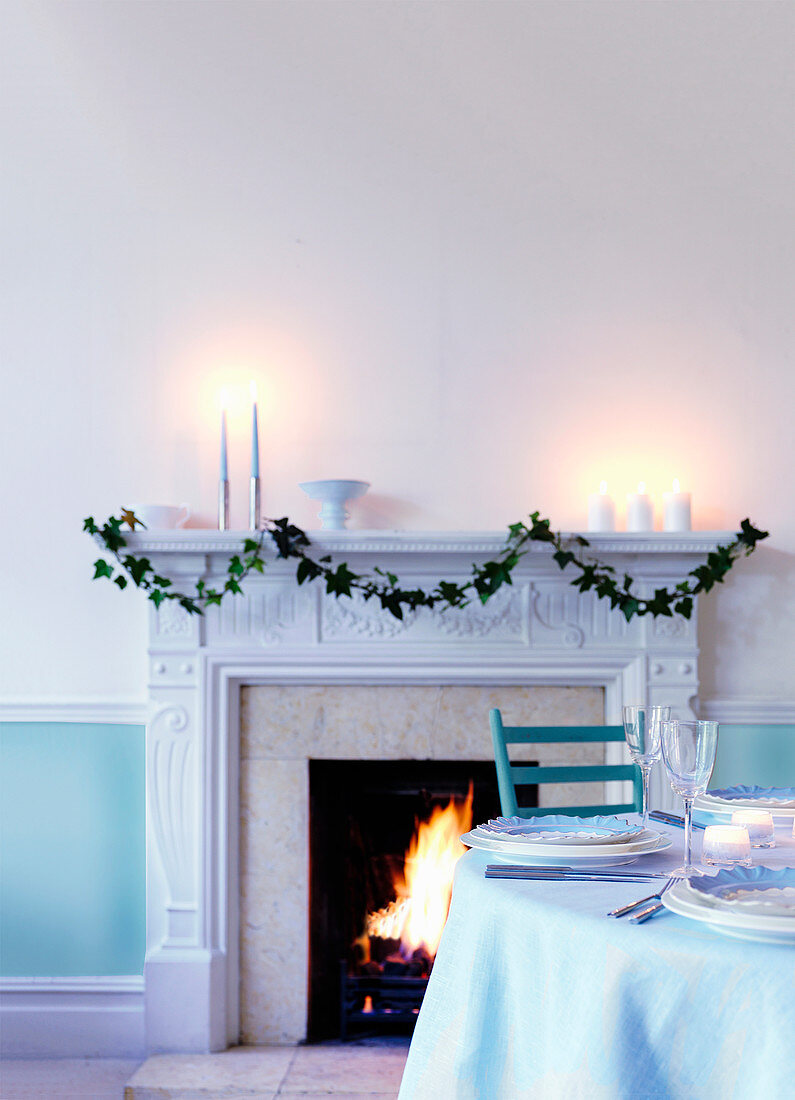 Gedeckter Tisch in kühlem Blau-Weiß am Kamin mit Efeuranke und brennenden Kerzen