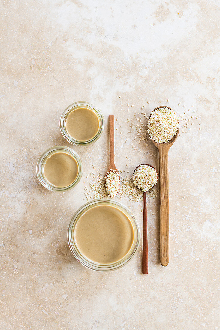 Tahini in jars next to sesame seeds on spoons (seen from above)
