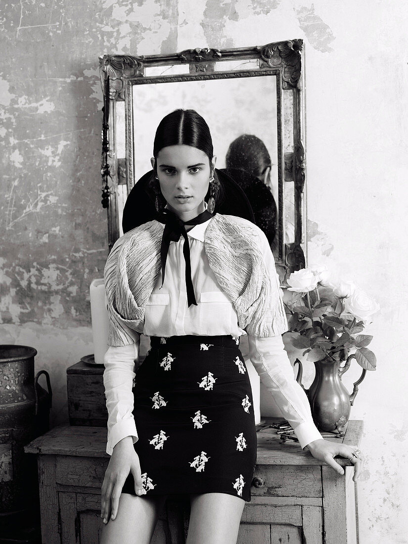 A young brunette woman wearing a Spanish outfit with a hat leaning against a chest of drawers