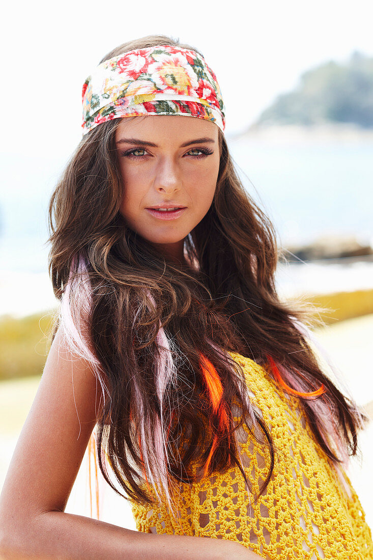 A young brunette woman with hair extensions by the sea wearing a yellow knitted top and a headscarf