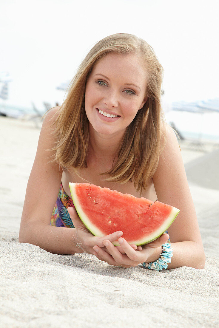 A young blonde woman on a beach wearing a colourful summer dress holding a wedge of melon