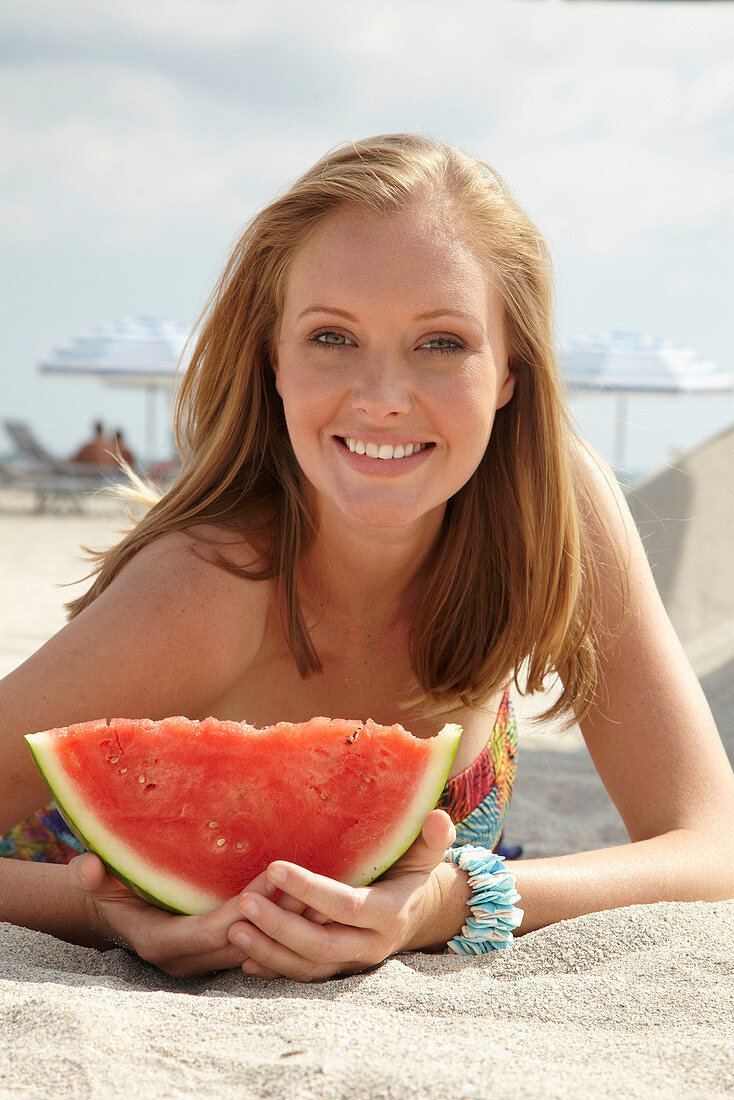 Junge blonde Frau im bunten Sommerkleid hält Melone am Strand