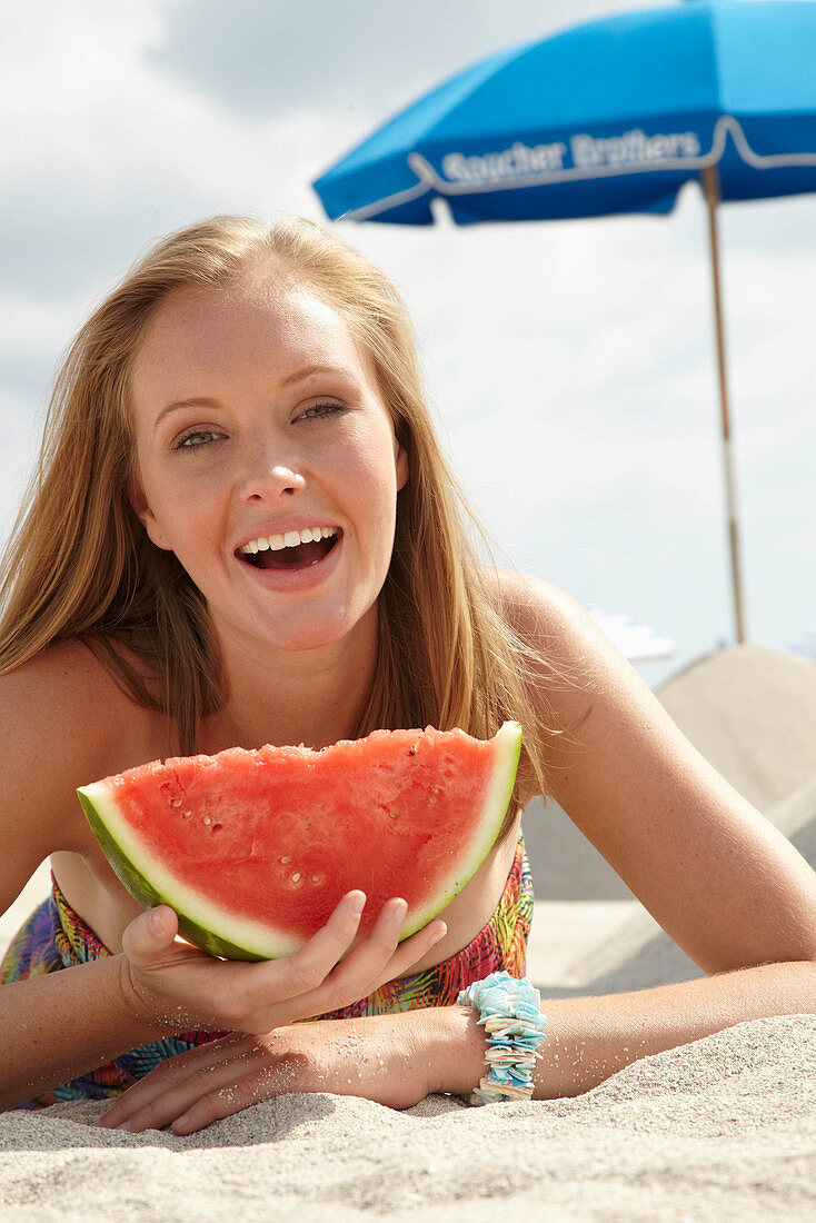 Junge blonde Frau im bunten Sommerkleid hält Melone am Strand