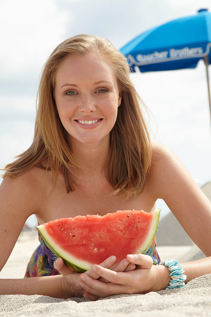 Junge blonde Frau im bunten Sommerkleid hält Melone am Strand