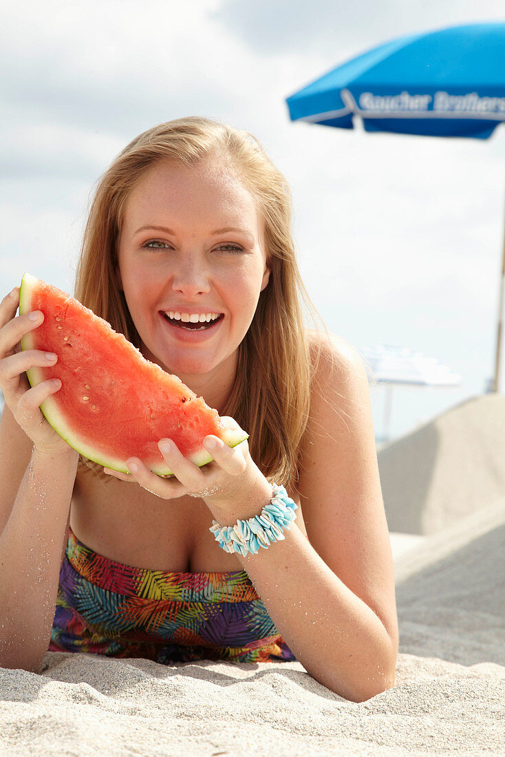 A young blonde woman on a beach wearing a colourful summer dress holding a wedge of melon