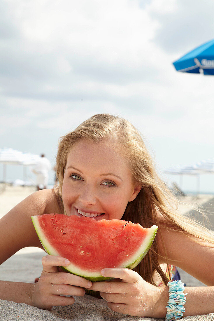 Junge blonde Frau im bunten Sommerkleid hält Melone am Strand