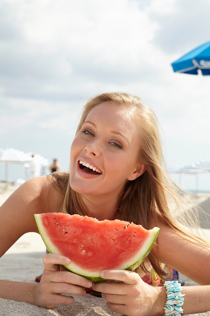 Junge blonde Frau im bunten Sommerkleid hält Melone am Strand