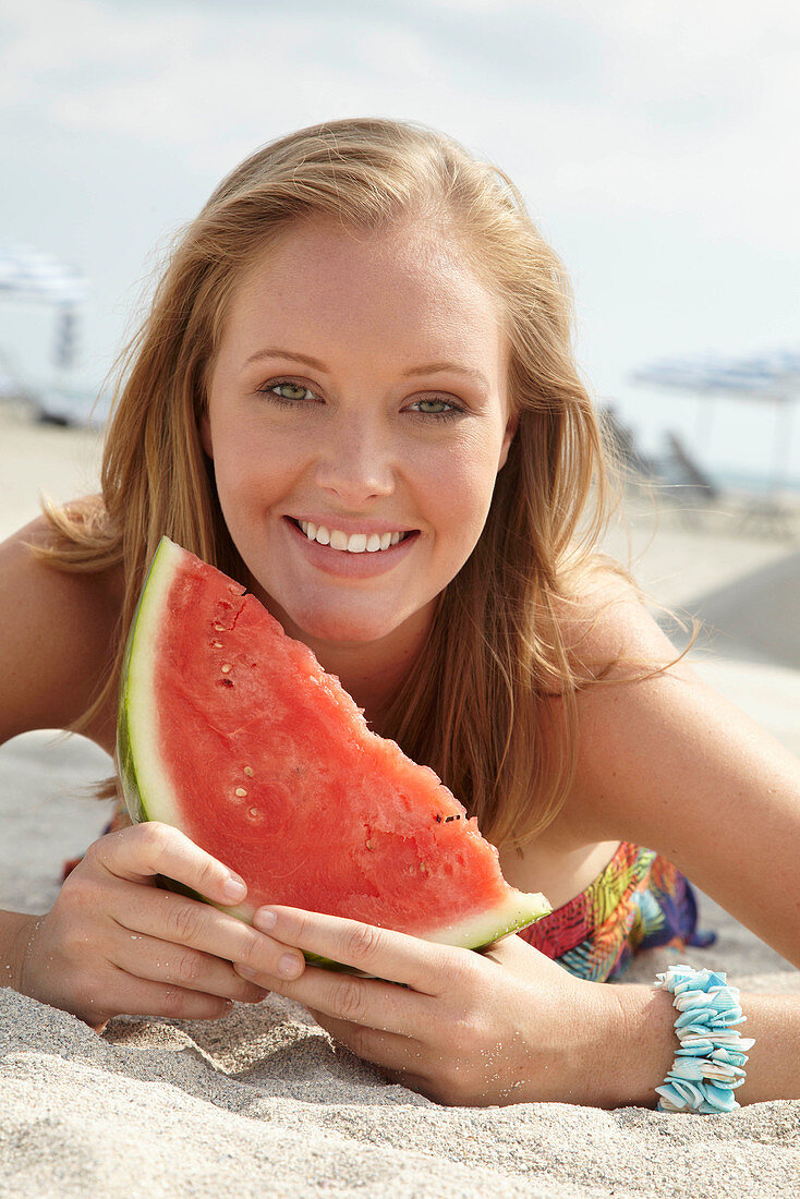 Junge blonde Frau im bunten Sommerkleid hält Melone am Strand