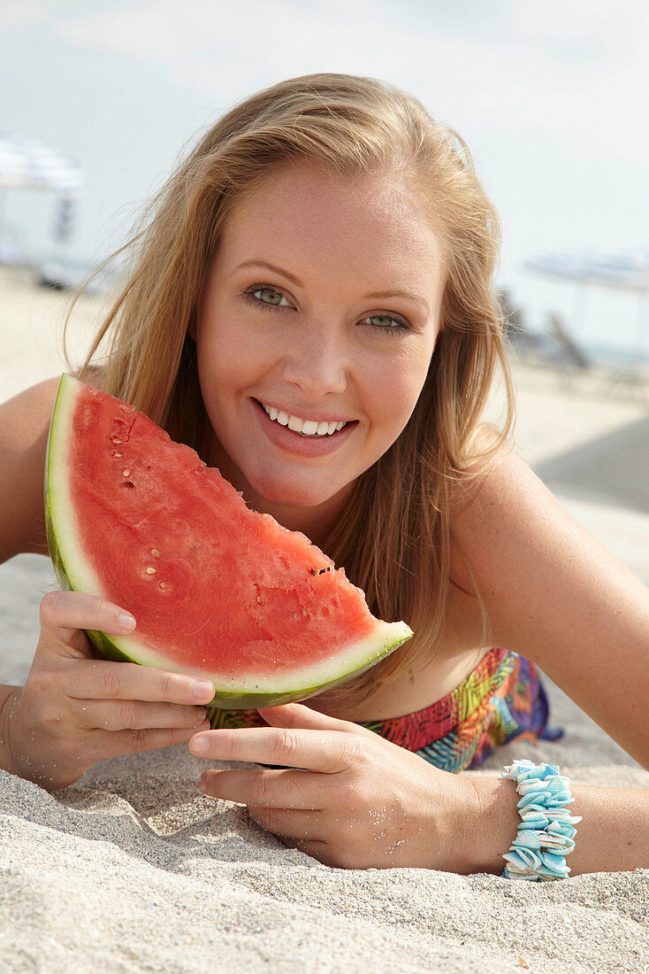 Junge blonde Frau im bunten Sommerkleid hält Melone am Strand