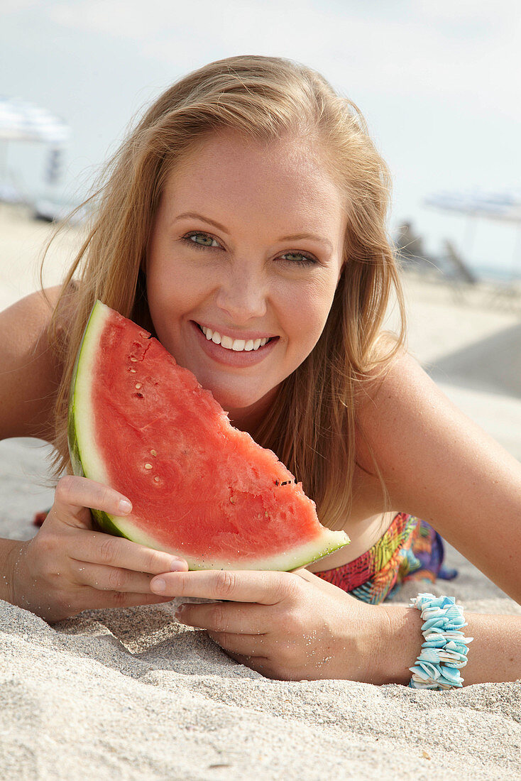 Junge blonde Frau im bunten Sommerkleid hält Melone am Strand