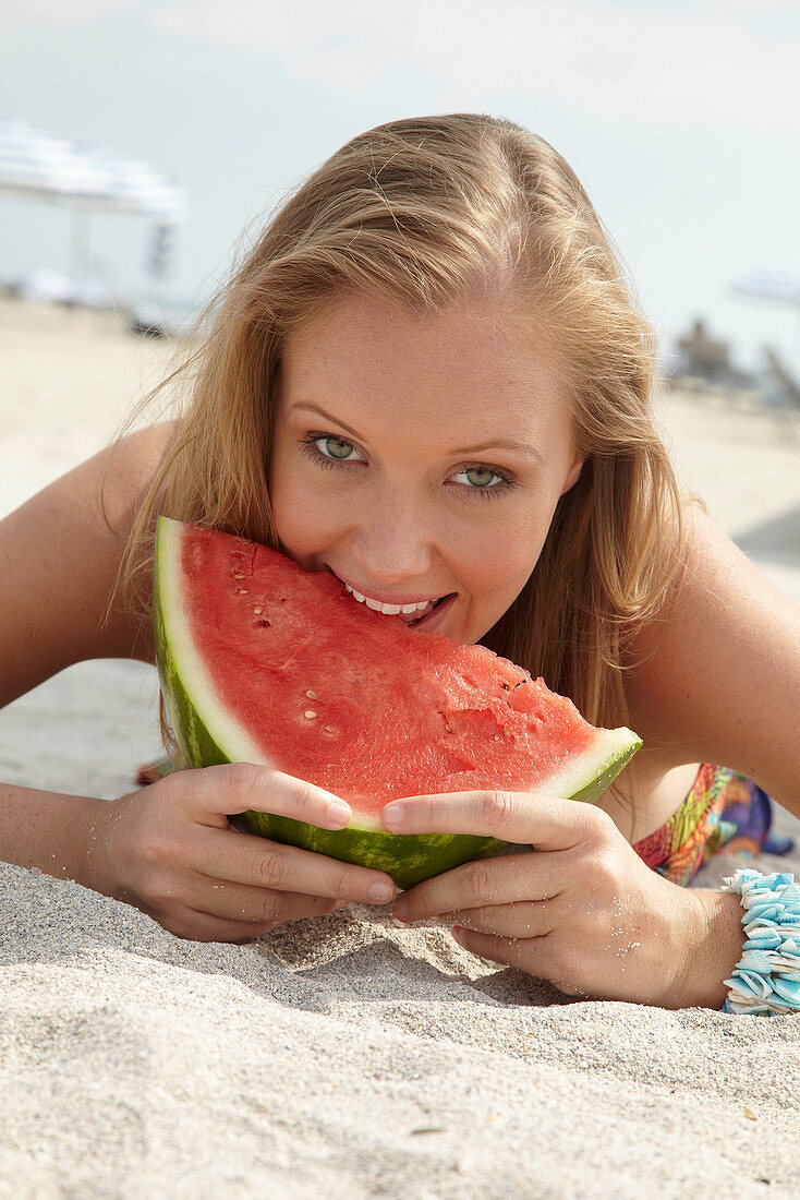 A young blonde woman on a beach wearing a colourful summer dress holding a wedge of melon