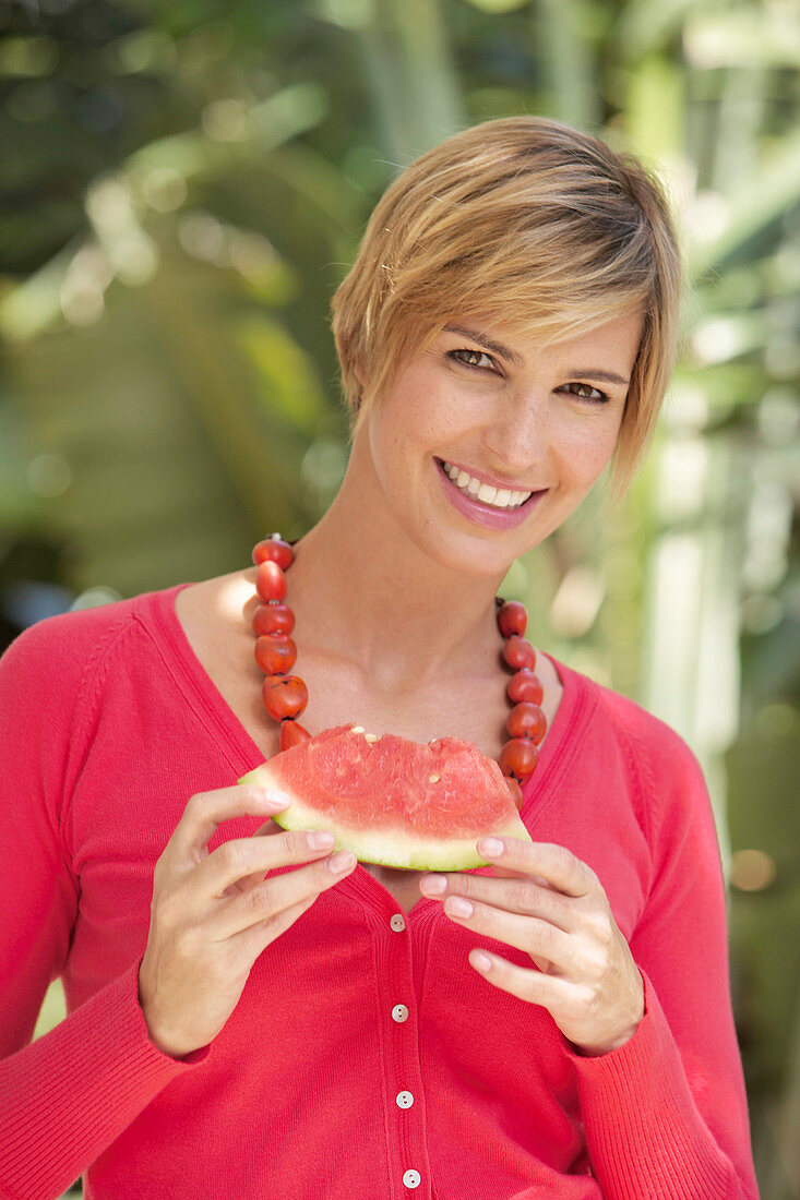A mature woman with short blonde hair outside wearing a red shirt and holding a slice of watermelon