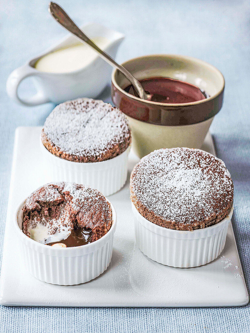 Three chocolate souffles dusted with icing sugar with bowl of melted chocolate and jug of pouring cream