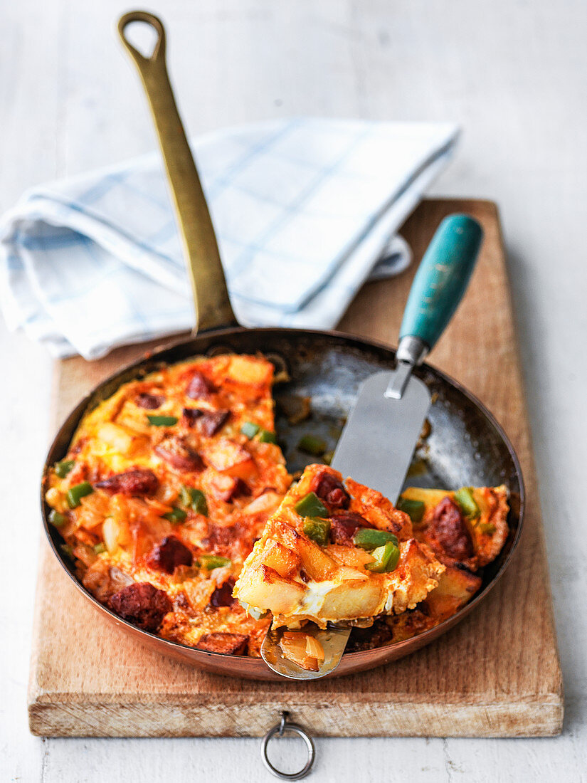 Spanish tortilla in fry pan with chorizo green peppers potatoes onions pumpkin and eggs showing slices out on pallette knife