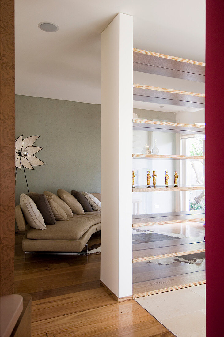 Sandy, curved sofa with scatter cushions against grey wall with partition wall in foreground