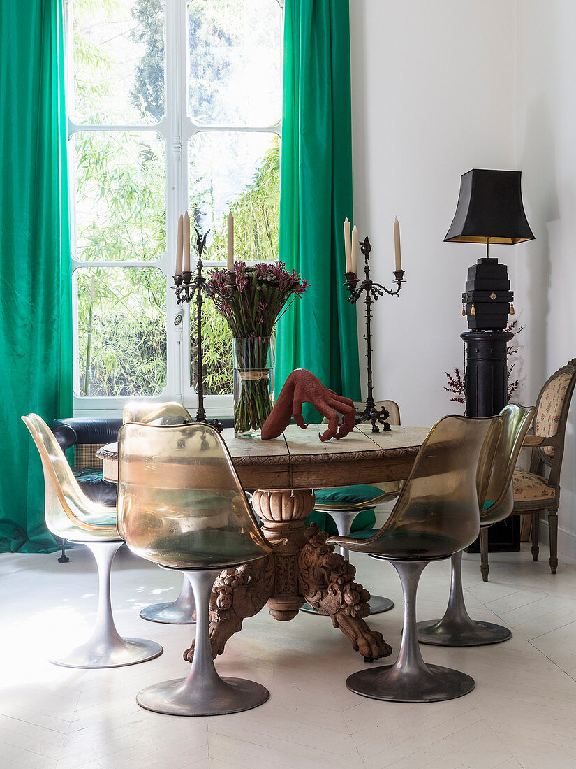 Runder Holztisch mit geschnitztem Tischbein und Klassikerstühle vor Fenster mit grünem Vorhang