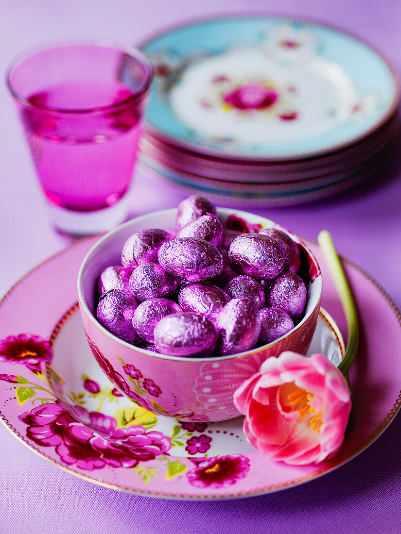 Schale mit Schokoladen-Ostereier in lila Folie und eine Tulpe auf geblümtem Teller
