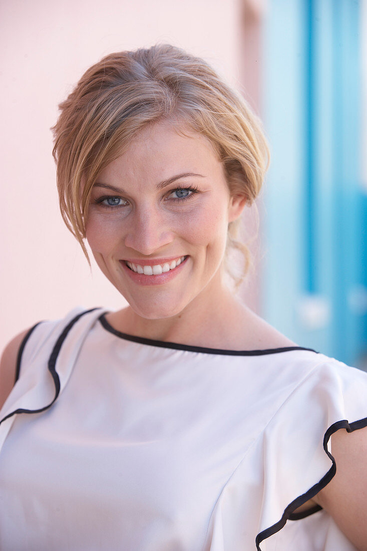A blonde woman standing against a pink background wearing a light blouse with a black trim