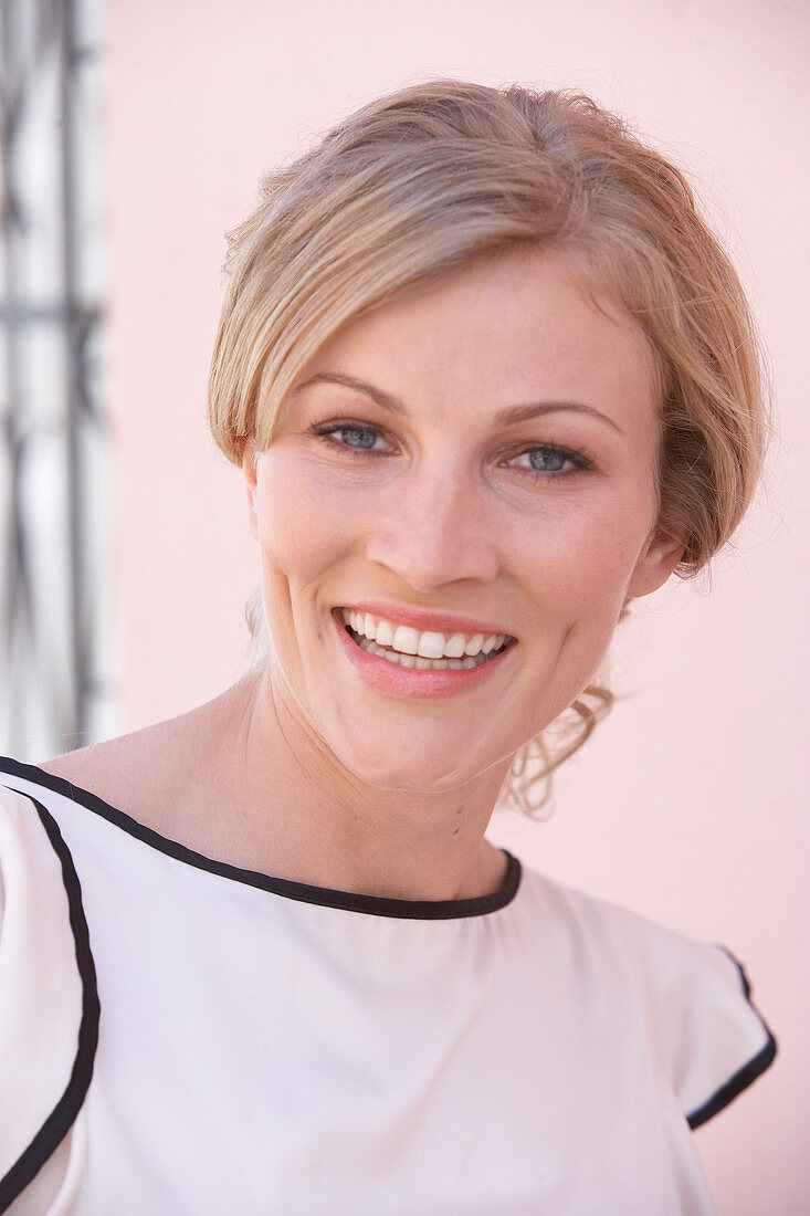 A blonde woman standing against a pink background wearing a light blouse with a black trim