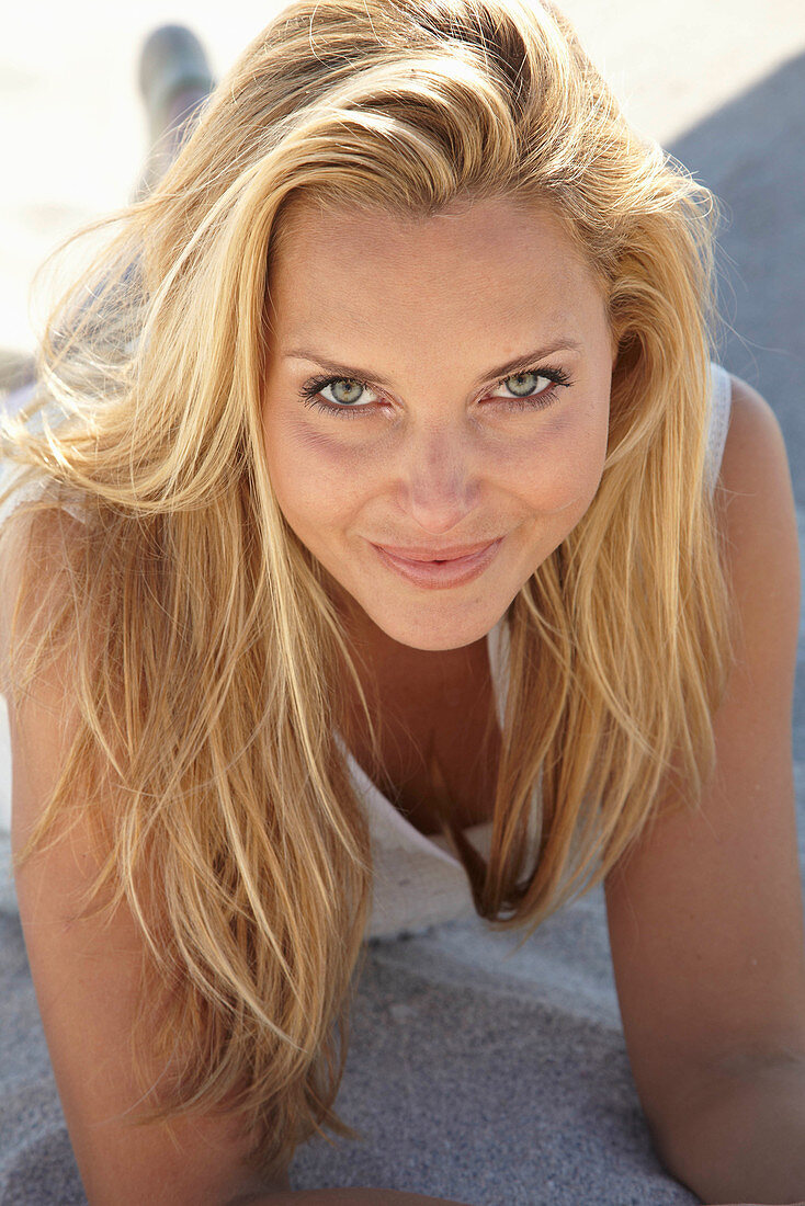 A mature blonde woman lying on a sandy beach wearing a white top