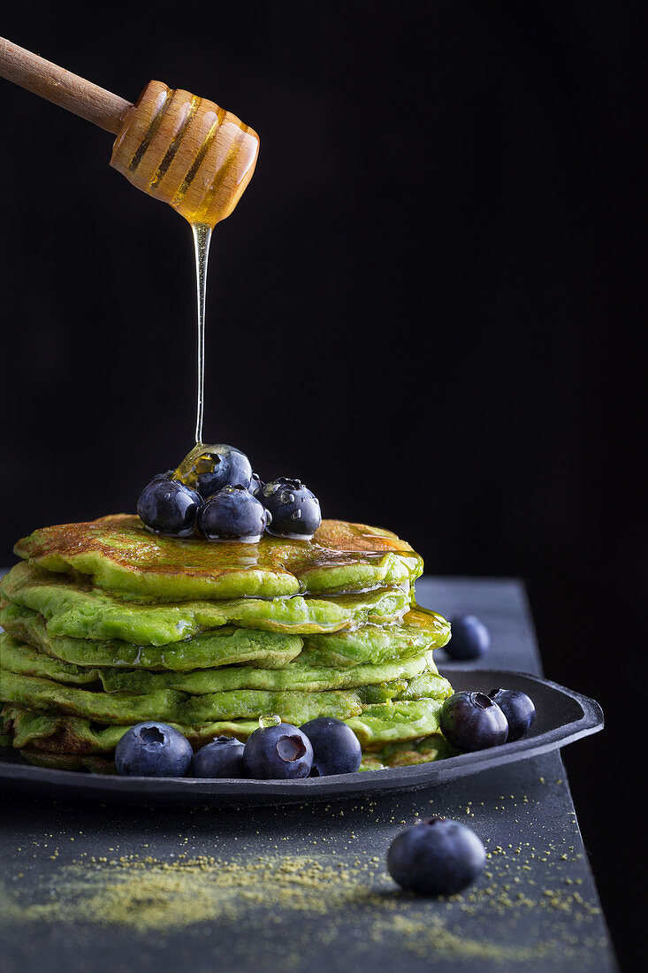 Honey pouring from wooden spoon to tasty green pancakes with blueberry