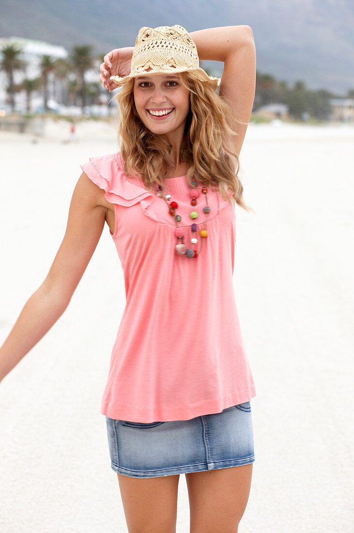 A young blonde woman on a beach wearing a pink top, a short denim skirt and a beige hat