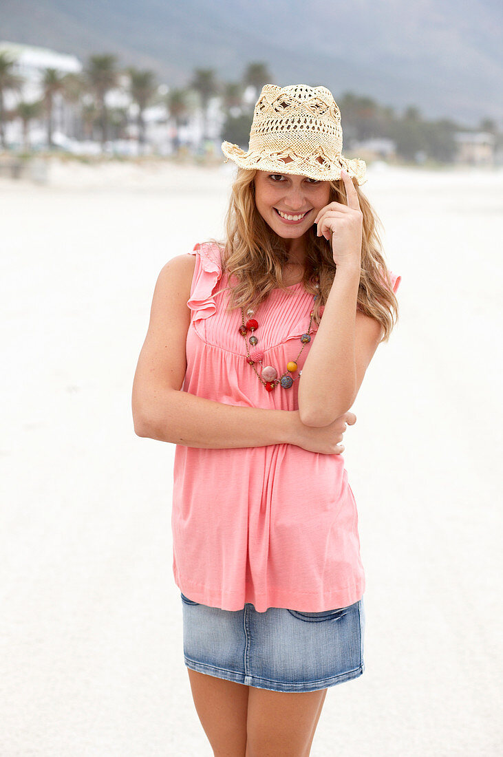 Junge blonde Frau im rosa Top, kurzem Jeansrock und beigem Hut am Strand
