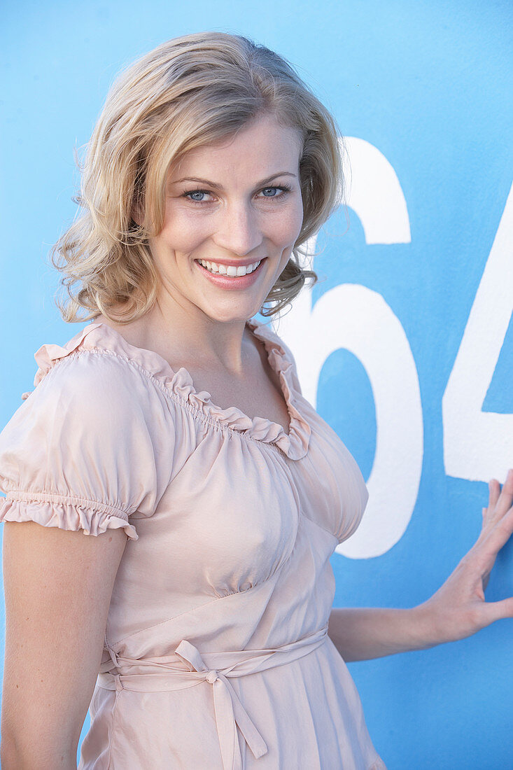 A blonde woman standing against a blue surface wearing a pink short-sleeved blouse