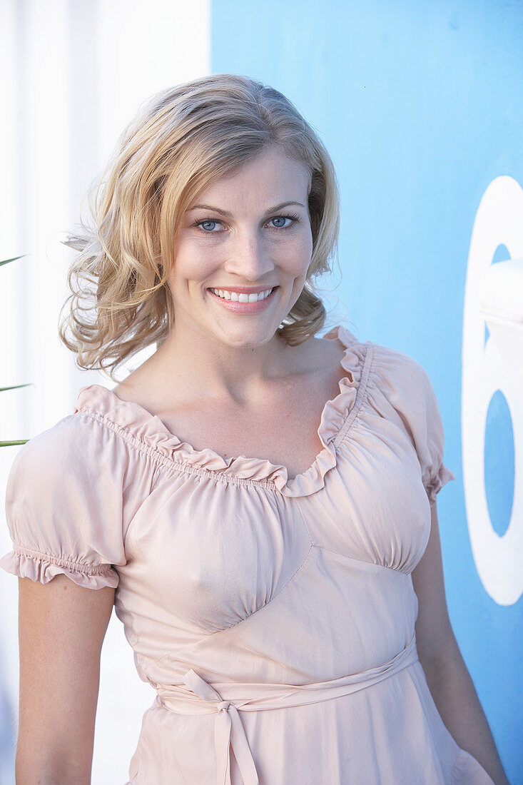 A blonde woman standing against a blue surface wearing a pink short-sleeved blouse