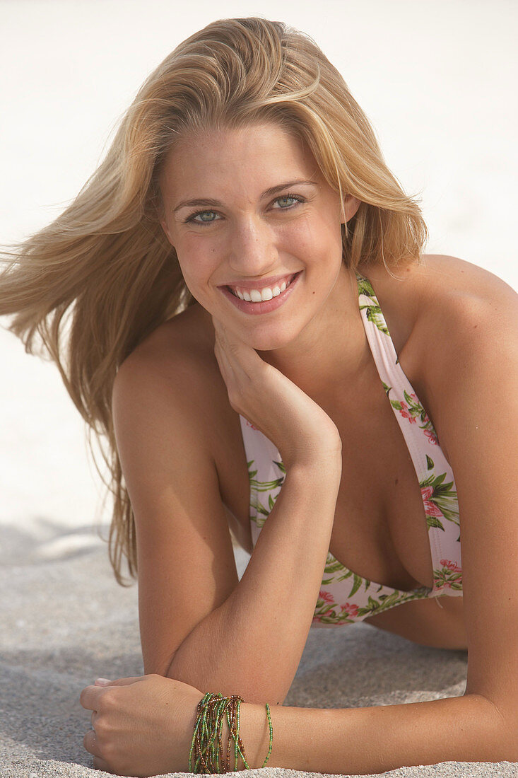 Junge blonde Frau im gemusterten Bikini liegt am Sandstrand