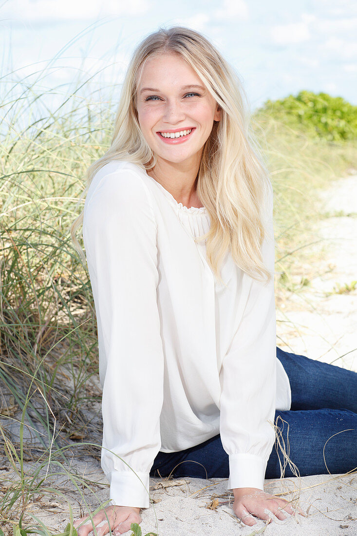 A young brunette woman sitting in the sand wearing a white blouse and jeans