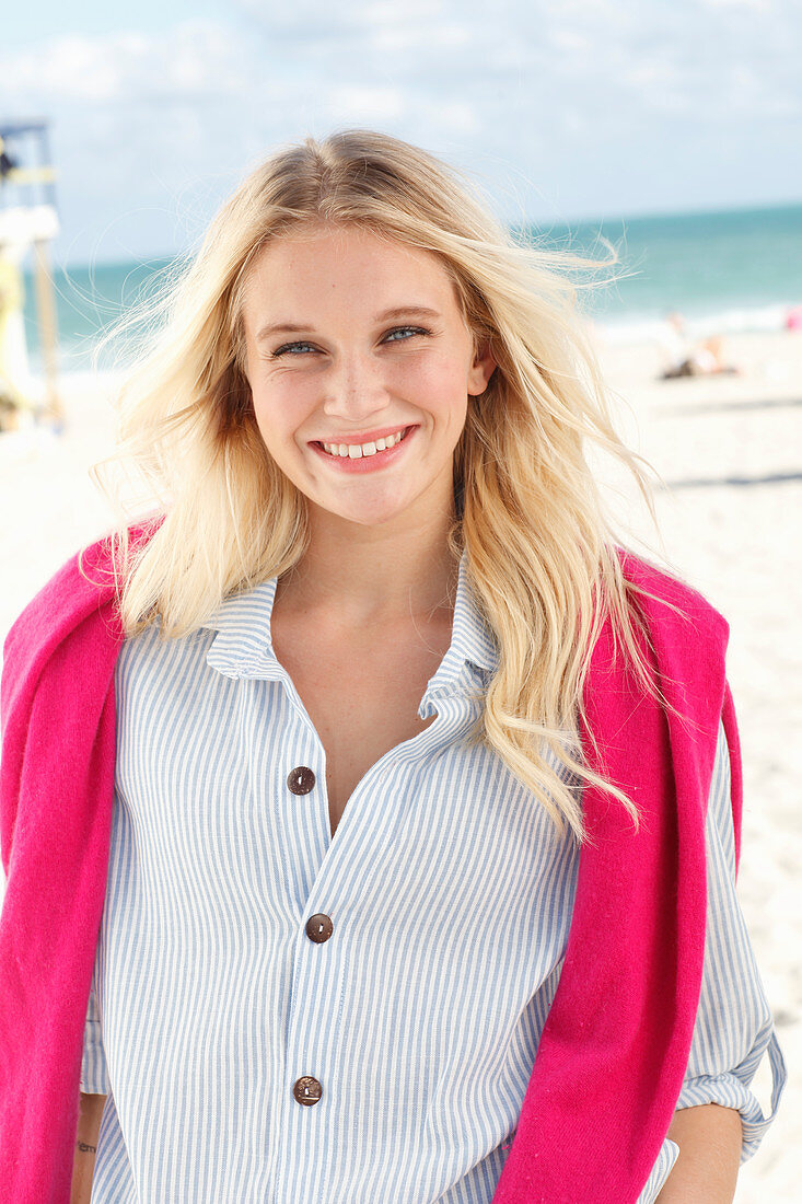 Junge blonde Frau in gestreiftem Hemd und mit pinkfarbenem Pulli am Strand